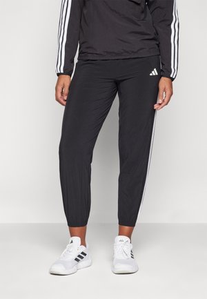 Persona que lleva pantalones deportivos negros de Adidas con rayas blancas y zapatillas Adidas blancas a juego, de pie frente a un fondo liso.