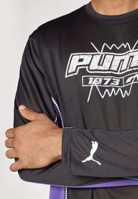 Μαύρη αθλητική μπλούζα με μακρύ μανίκι, που διαθέτει το λογότυπο "PUMA" σε λευκό χρώμα, υφή υφάσματος, μωβ λεπτομέρεια στο στρίφωμα και έμβλημα με λευκή πάνθηρα στο μανίκι.
