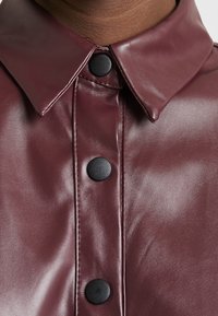 Burgunderfarbenes Kunstlederhemd mit Spitzkragen, ausgestattet mit schwarzen runden Knöpfen und einer glatten, glänzenden Textur.