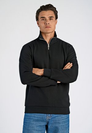Sudadera negra con cremallera en el cuello y mangas largas, hecha de una tela suave. Presenta una cremallera plateada y un corte relajado, combinada con jeans azules.