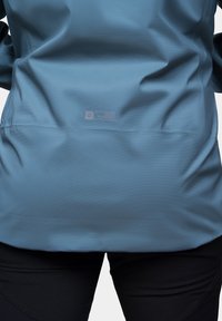 Wodoodporna niebieska kurtka z teksturowanej tkaniny, z metką z tyłu, zestawiona z czarnymi spodniami.