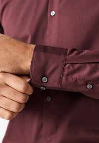 Camicia da uomo color bordeaux con tessuto strutturato, leggero lucido e bottoni grigi su polsino e patta; dal design aderente.