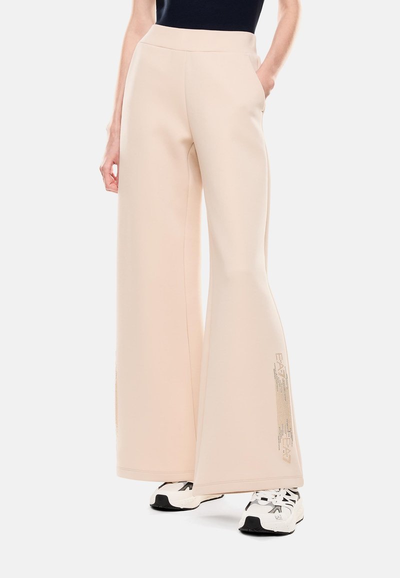 Pantalones de pierna ancha en color beige con una textura suave y cintura alta, que presentan bolsillos laterales y un sutil detalle del logo en la pierna izquierda.