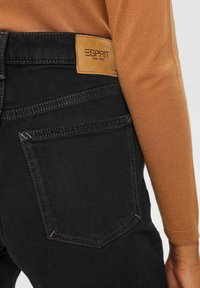 Gros plan sur une personne portant un jean noir avec une étiquette en cuir Esprit New York et un haut à manches longues marron.