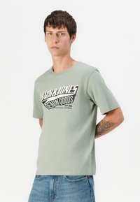 Lys grøn bomulds T-shirt med rund hals, der har et stort sort-hvidt grafisk tryk, korte ærmer og lige kant.