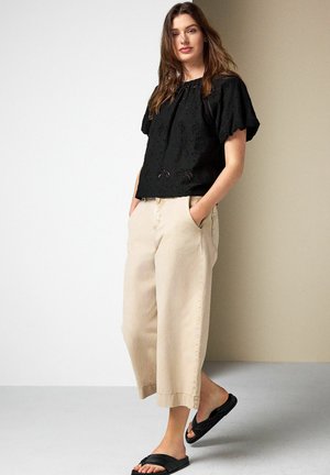 Woman wearing black eyelet short-sleeve blouse, beige wide-leg cropped pants, and black slide sandals, standing with hands in pockets.