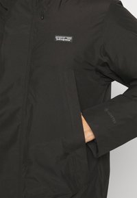Czarna kurtka z Gore-Texu z zamkiem błyskawicznym, przestronnymi kieszeniami i logo Patagonia. Gładka tekstura o lekko błyszczącym wykończeniu.