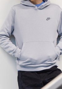 Šedá bavlněná mikina s kapucí, přední kapsou a malým černým logem Nike na hrudi. Nositá přes modré tričko.