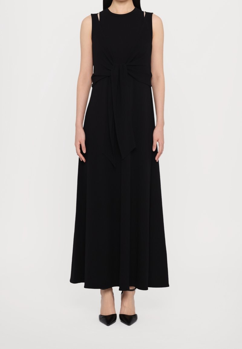 Femme portant une robe maxi noire sans manches avec un nœud à l'avant et des talons noirs à bout pointu, debout sur fond blanc.