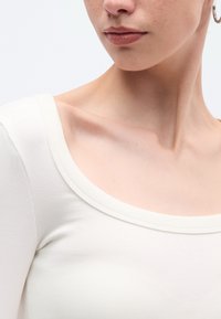 Λευκό ριμπ πλεκτό μπλουζάκι με μακριά μανίκια και φαρδύ, στρογγυλεμένο λαιμόκομμα. Ομαλή υφή, εφαρμοστό σχέδιο και απαλό ύφασμα, αναδεικνύοντας την κλείδα.