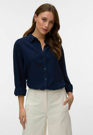 Femme aux longs cheveux ondulés portant une chemise bleu marine boutonnée avec des manches retroussées et un pantalon crème taille haute, debout devant un fond uni.
