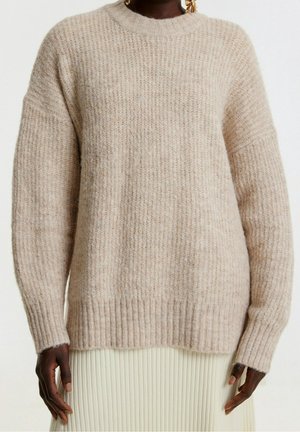 Maglione beige lavorato a maglia con texture a costine, maniche ampie e scollatura a girocollo. Materiale morbido e vestibilità rilassata. Sotto il vestito c'è una gonna plissettata di colore chiaro.