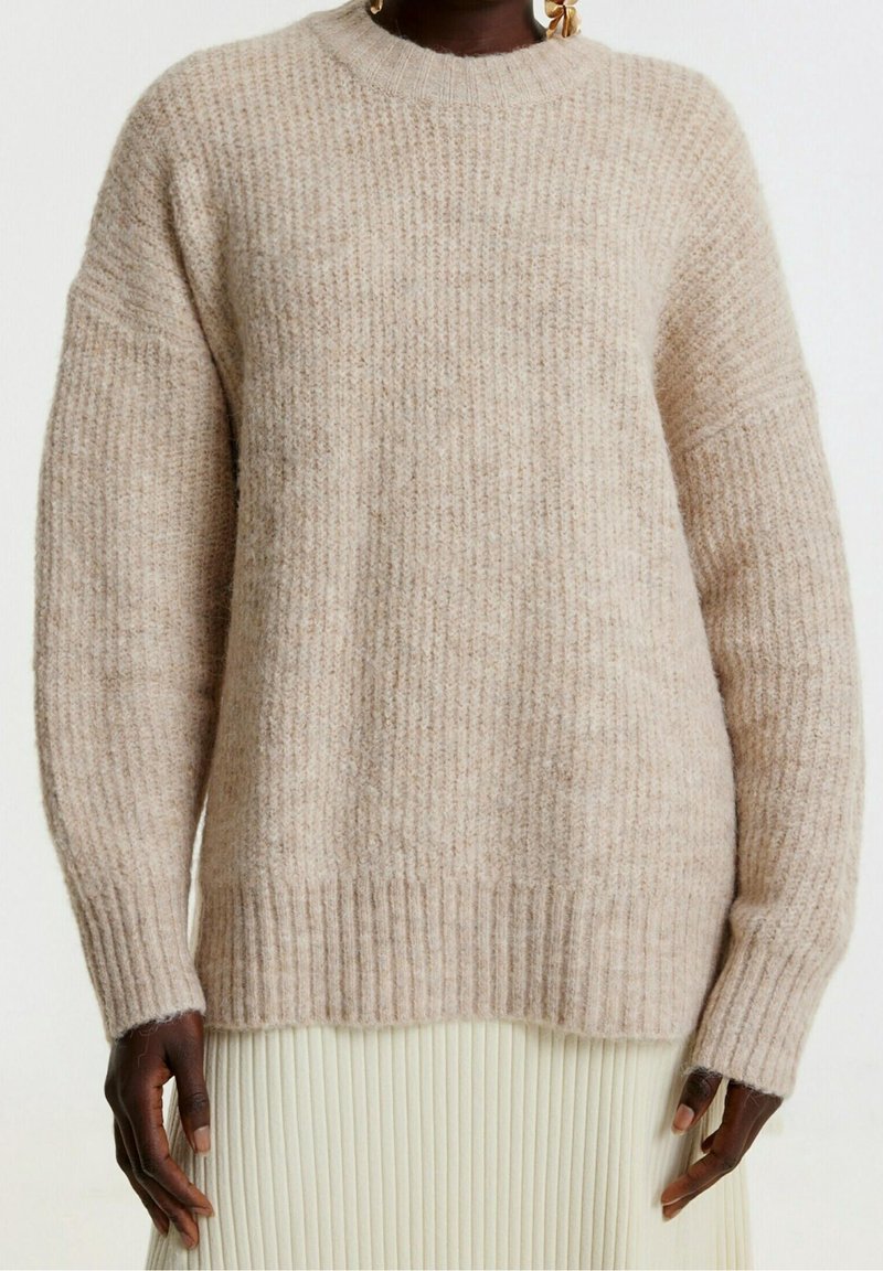 Maglione beige lavorato a maglia con texture a costine, maniche ampie e scollatura a girocollo. Materiale morbido e vestibilità rilassata. Sotto il vestito c'è una gonna plissettata di colore chiaro.