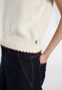 Suéter sin mangas color crema con ribete acanalado y acento de color burdeos oscuro. Textura suave, combinado con jeans de mezclilla azul oscuro.