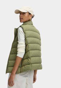 Groene, puffer vest met horizontale stiksels, hoge kraag en rits. Gecombineerd met een crèmekleurige trui en een licht pet, staat het model met de rug naar de camera gekeerd.