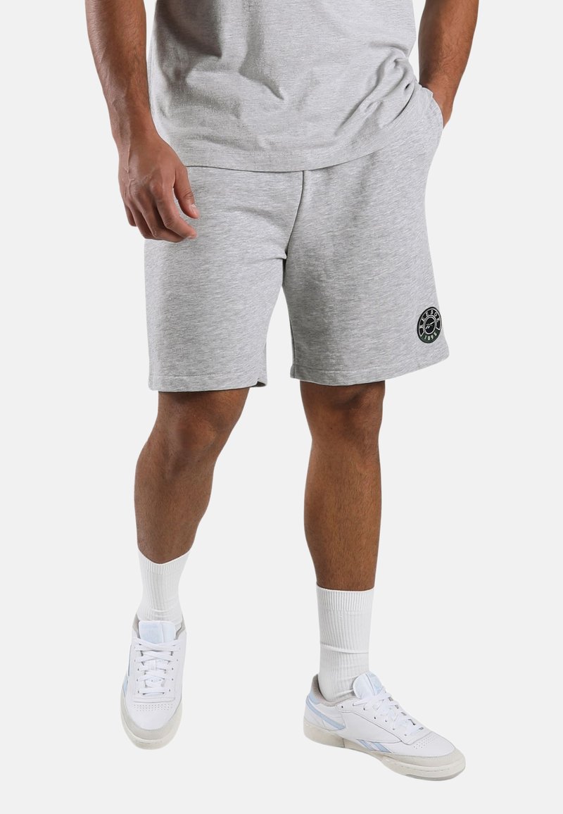 Man wearing light gray athletic shorts with logo on left leg, white crew socks, and white sneakers, standing against a plain background.