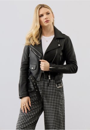 Blonde Frau trägt schwarze Leder-Bikerjacke, weißes Oberteil, silbernen Schmuck und schwarz-weiß karierte Hose, posiert vor schlichtem Hintergrund.