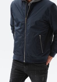 Marineblå bombarjacka med dragkedja fram, ribbade ärmslut och nederkant, med två sidfickor med dragkedja och en grå huva som tittar fram från kragen.