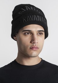 Beanie nero a maglia con lettering in rilievo "Kavanagh" e "Gianni" sul risvolto. Tessuto testurizzato e elastico che si adatta alla testa.