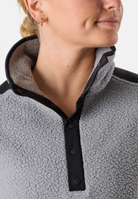 Grå fleece-tröja med svart krage och knappstängning. Har en texturerad yta och en hög hals för värme.