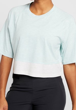 Person, die ein hellblaues, kurzes T-Shirt mit weißem Saum und dunkle, hoch taillierte, eng anliegende Hosen trägt, vor einfarbigem Hintergrund.