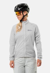 Lichtgrijs windjack met hoge kraag en frontale rits, met het Jack Wolfskin-logo. Draagt een zwarte fietshelm en een zonnebril.