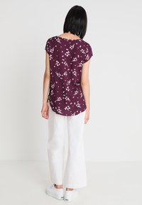 Kobieta z krótkimi ciemnymi włosami, ubrana w fioletową kwiatową bluzkę z krótkim rękawem, białe szerokie spodnie i białe trampki, odwrócona plecami.