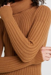 Pull à col roulé côtelé marron avec des manches longues, présentant un col épais et un tricot texturé, mettant en avant des lignes verticales et un aspect doux et confortable.