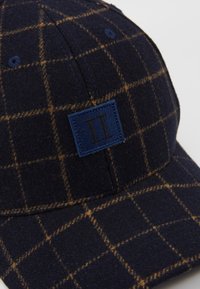 Casquette à carreaux bleu marine avec un motif brun à carreaux et un patch bleu marine affichant deux lignes noires verticales sur le panneau avant.