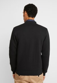 Mand iført en sort tekstureret jakke over en blå skjorte med krave og beige bukser, vist fra ryggen mod en ensfarvet baggrund.