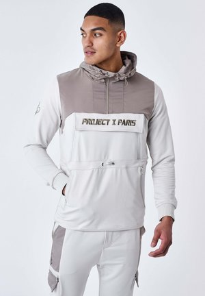 Hombre con sudadera con capucha de color gris claro y topo y pantalones a juego con el texto "Project X Paris" en el pecho, mirando hacia la izquierda con las manos parcialmente en los bolsillos.