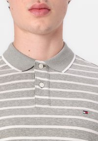 Γκρι και λευκή ριγέ πόλο φανέλα φτιαγμένη από υφασμάτινη υφή, με κουμπωτό γιακά και ένα μικρό λογότυπο στην αριστερή πλευρά.