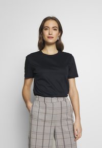 Schwarzes kurzes T-Shirt aus glattem Stoff, kombiniert mit grauen karierten Hosen mit braunen und marineblauen Linien, ausgestattet mit seitlichen Taschen.