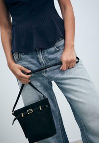 Persona che indossa una maglia a righe scure e jeans chiari, mentre tiene una borsa a secchiello nera con una fibbia dorata per la sua lunga tracolla.