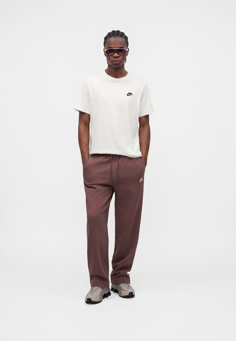 Junger Mann mit geflochtenem Haar, trägt dunkle Sonnenbrille, weißes Nike-T-Shirt, mauvefarbene Nike-Sweathose und beige Sneakers, steht mit den Händen in den Taschen.