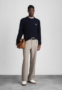 Mornarsko pleteno džemper s malim logom, bež uzorčane hlače i krem bijele tenisice. Drži smeđi ruksak. Jednostavan, čist dizajn.