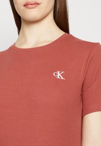 Žebrované tričko s krátkým rukávem v barvě cihlové, s kulatým výstřihem. Na levém prsu se nachází malé bílé logo "cK". Materiál je měkká směs bavlny.