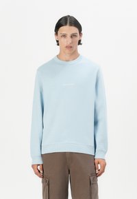Lyseblå sweatshirt med ribbet rundt hals og manchetter. Har et diskret hvidt logo på brystet og en afslappet pasform. Kombination med brune cargo-bukser.