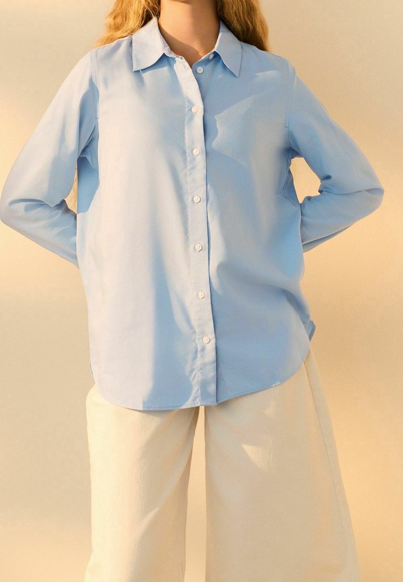 Camicia azzurro chiaro con bottoni realizzata in un tessuto morbido, con colletto, maniche lunghe e orlo curvo, abbinata a pantaloni bianchi a gamba larga.