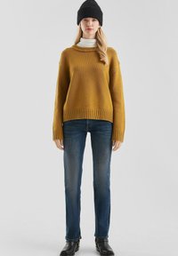 Musztardowy, oversize'owy sweter dzianinowy z prążkowanymi mankietami, w połączeniu z ciemnoniebieskimi spodniami typu straight-leg i czarną dzianinową czapką.