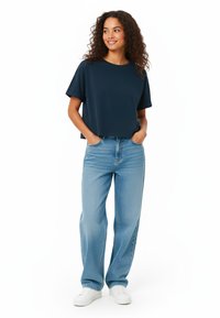 Mornarsko modra, kratka majica s kratkimi rokavi v kombinaciji z svetlimi, visokim pasom jeans hlačami in belimi športnimi copati. Model stoji z rokama v žepih.