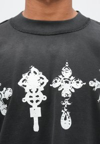 Fekete pamut póló texturált, kopott fehér díszítőmintákkal a mellkas közepén. Kerek nyakú kialakítás, látható varrással.
