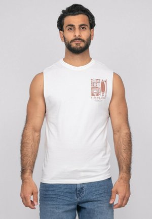 Homme barbu portant un débardeur blanc avec un motif marron et un jean bleu, debout devant un fond clair uni.