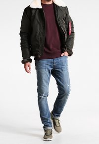 Dunkelgrüne Bomberjacke mit weißem Pelzkragen, kombiniert mit einem kastanienbraunen Sweatshirt, hellblauen zerrissenen Jeans und grauen Sportschuhen.