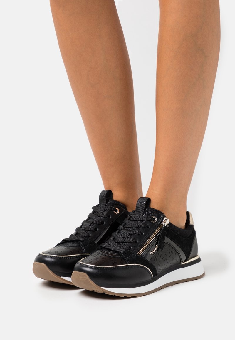Sneakers bassi neri e marroni con lacci e zip laterale indossati su gambe nude su uno sfondo bianco.