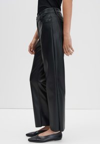 Persoon in zwarte wijde neplederen broek en zwarte puntige platte schoenen, staand in profiel tegen een effen lichte achtergrond.