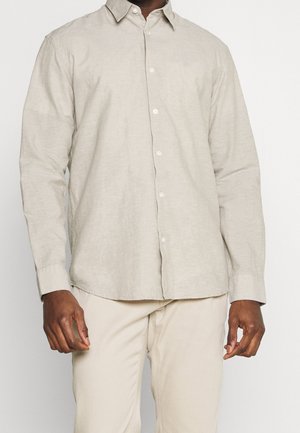 Homme portant une chemise beige clair à manches longues boutonnée et un pantalon beige, debout devant un fond uni.