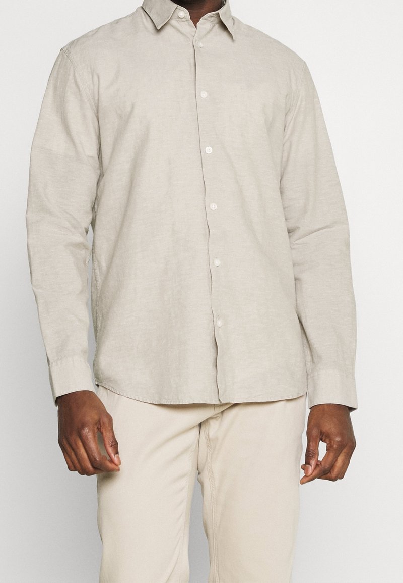 Homme portant une chemise beige clair à manches longues boutonnée et un pantalon beige, debout devant un fond uni.