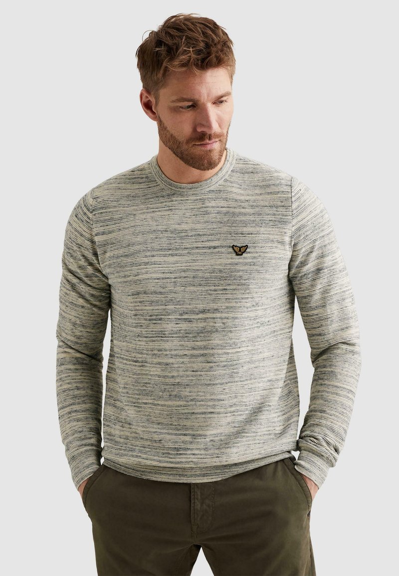 Homme portant un pull tricoté texturé gris clair avec un petit emblème d'aigle noir et or sur la poitrine, les mains dans les poches d'un pantalon vert foncé.