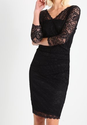 Femme portant une robe noire en dentelle longueur genou avec des manches trois-quarts, debout avec un bras croisé et la main près du visage.